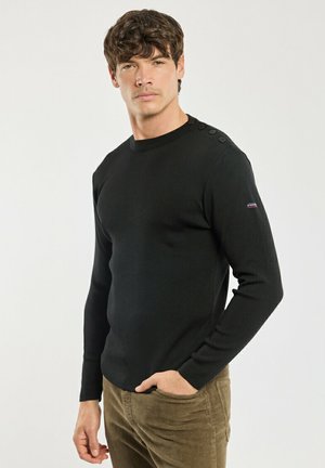 Mand iført en sort langærmet sweater med knapper på skulderen og brune kordbukser, står med den ene hånd i lommen.