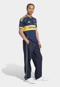 Tamsiai mėlyna futbolo marškinėliai su geltonais akcentais ir logotipu, derinami su tamsiomis džinsinio audinio kelnių su šoninėmis juostelėmis ir baltais sportbačiais.