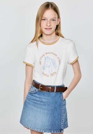 Dziewczyna z długimi blond włosami, ubrana w białą koszulkę z grafiką konia, perłowy naszyjnik, brązowy pasek i jeansową spódnicę, z ręką w kieszeni.