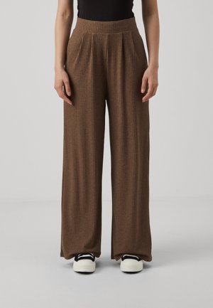 Vrouw met bruine broek met brede pijpen en ribstructuur, gecombineerd met een zwart topje en witte sneakers, staand tegen een effen lichte achtergrond.