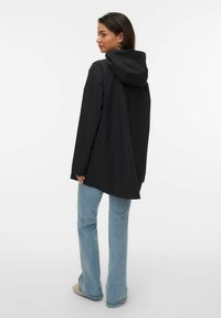 Veste noire à capuche à coupe ample, fabriquée en matériau doux, avec une longueur mi-cuisse. Associée à un jean bleu clair évasé.