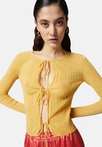 Top giallo a costine con scollatura a V profonda e chiusura con nodo davanti. Maniche lunghe, texture leggera e silhouette aderente.