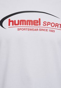 Witte stof met rode tekst "hummel SPORT" en "SPORTSWEAR SINCE 1923" onder een zwarte gebogen lijn.