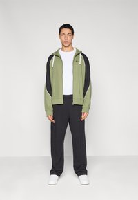 Sweat à capuche zippé vert et noir avec des accents blancs, associé à un pantalon large gris foncé et des baskets blanches. Le mannequin se tient devant un fond clair.
