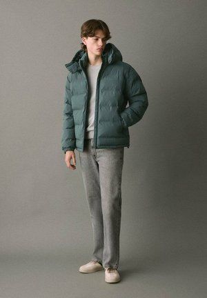 Giacca a vento verde con cappuccio, chiusura con zip e polsini a costine, abbinata a jeans grigi chiari e sneakers beige su uno sfondo neutro.