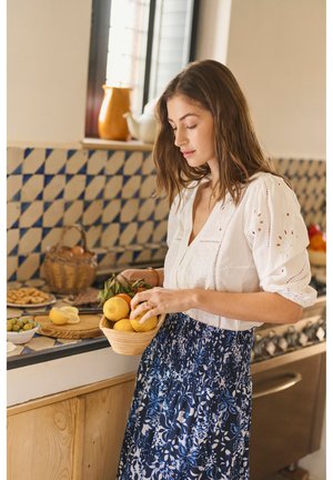 Noor naine valges pluusis ja sinises seelikus hoiab plaaditud köögis sidrunikorvi ja valmistab toitu.