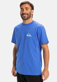 Modrá bavlnená tričko s okrúhlym výstrihom, krátkymi rukávmi a malým svetlomodrým logom na hrudi; nosené s čiernymi nohavicami.