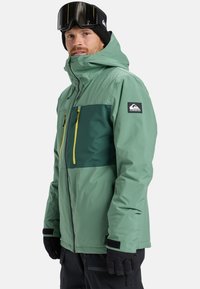 Grüne und dunkelblau-teal wasserfeste Jacke mit gelben Reißverschluss-Details, ausgestattet mit einem hohen Kragen und mehreren Reißverschlusstaschen. Schwarze Handschuhe und Skibrille.
