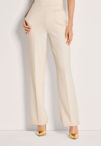 Vrouw die een hooggetailleerde crèmekleurige broek met voornaaien en gouden puntige hakken draagt, staande tegen een effen lichte achtergrond.