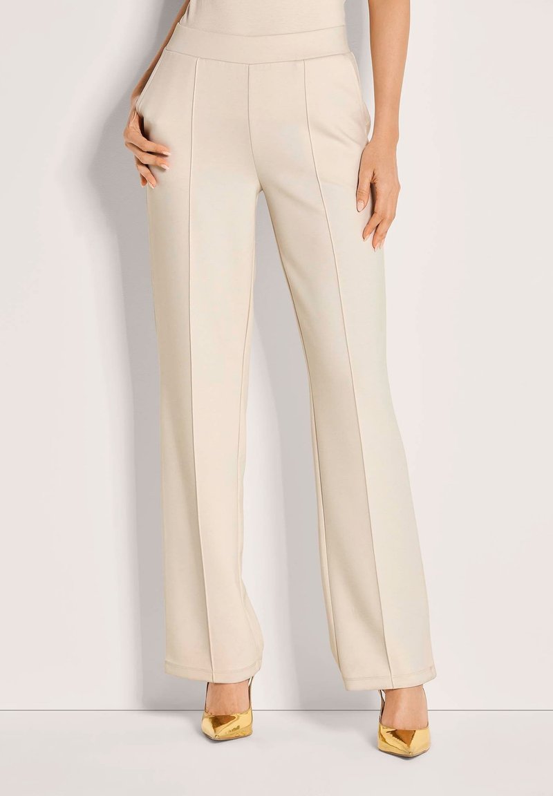 Vrouw die een hooggetailleerde crèmekleurige broek met voornaaien en gouden puntige hakken draagt, staande tegen een effen lichte achtergrond.