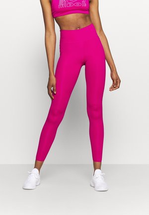 Femme portant des leggings taille haute rose vif, un crop top avec le texte « Just Do It » et des chaussures de sport blanches, se tenant devant un fond blanc.