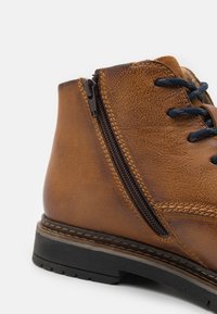 Brown leather ankle boot featuring a side zipper, black laces, textured surface, and a sturdy black rubber sole with stitch detailing.