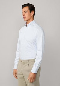 Muškarac u svijetloplavoj kariranoj košulji dugih rukava s dugmadi i bež hlačama, stoji naslonjen na praznu sivu pozadinu.