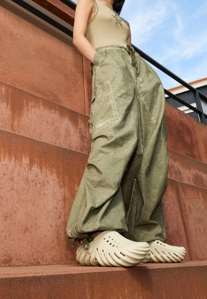 Persoon in oversized olijfgroene cargobroek, beige geribbelde tanktop en crèmekleurige sculpturale instappers die op roestkleurige treden staat.