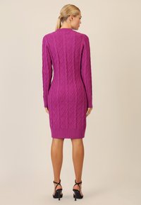 Fuchsia gebreide jurk met een gestructureerd diamanten patroon, lange mouwen, geribbelde zoom en een middellange lengte. Draagt een paar zwarte hoge hakken met banden.