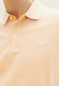 Pikétröja i ljus persika med en strukturerad tyg, kontrasterande vit randig krage och en diskret logotyp på bröstet.