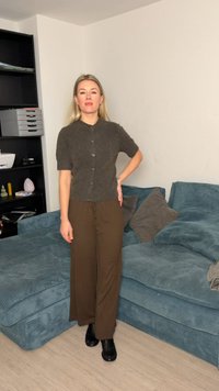 Ξανθιά γυναίκα στέκεται στο σαλόνι φορώντας καφέ παντελόνι, σκούρο πουκάμισο με κουμπιά και κοντά μανίκια και μαύρα παπούτσια δίπλα σε μια μπλε καναπέ από κορδόνι με μαξιλάρια.