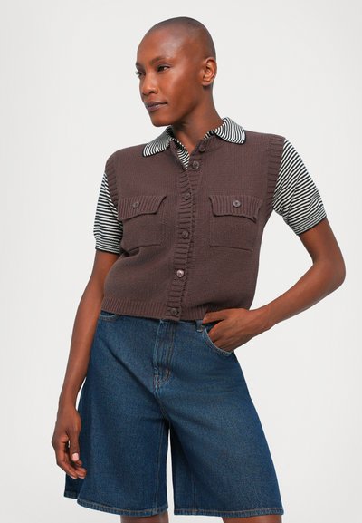 Gilet in maglia marrone con chiusura a bottone e due tasche frontali, indossato sopra una camicia a maniche corte a righe bianco e nera e pantaloni shorts di jeans.