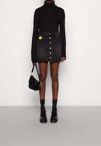 Maglione a collo alto nero abbinato a una gonna mini di denim nera con bottoni dorati e una tasca con un dettaglio giallo. Stivaletti neri e borsa.