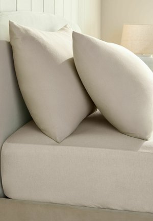 Two beige pillows leaning against a white headboard on a beige bed mattress with a lamp in the background.