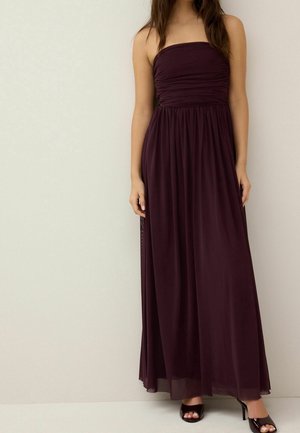 Vrouw in een strapless, vloerlange, donkerpaarse jurk en zwarte open teen hoge hakken, staand tegen een effen lichte achtergrond.