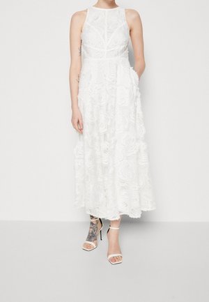 Kobieta ubrana w białą sukienkę midi z kwiatowej koronki bez rękawów z kieszeniami, nosząca białe sandały na wysokim obcasie z paskiem wokół kostki oraz widoczny tatuaż na nodze.