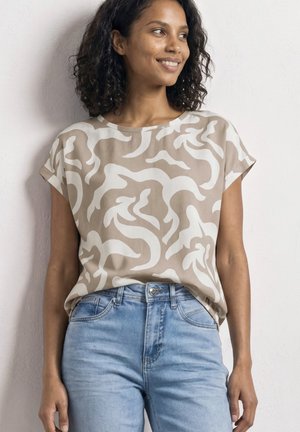 Femme aux cheveux bouclés portant un haut beige à manches courtes à motifs et un jean bleu clair, souriante et regardant sur le côté.