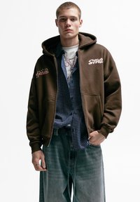 Brun lynlås-hættetrøje med store forlommer, hætte og broderede detaljer. Båret over en blå ternet skjorte og løstsiddende denimjeans.