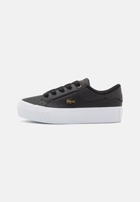 Zapatilla negra hecha de material sintético con suela de goma blanca. Presenta cordones redondos y un logotipo dorado en el lateral.