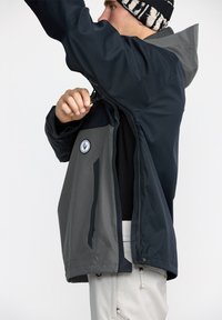 Veste imperméable bleu marine et grise avec fermeture éclair à l'avant, capuche ajustable et patch de logo. Le tissu semble lisse avec une finition mate.