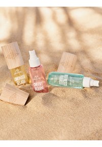 Tres botellas de spray con tapas de madera etiquetadas como Sol de Janeiro, en rosa, amarillo y turquesa, parcialmente enterradas en la arena de una playa iluminada por el sol.