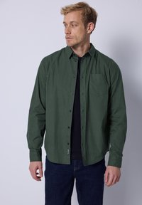 Camisa de botones de color verde oscuro con un patrón texturizado, un solo bolsillo en el pecho y botones negros, sobre una camiseta negra. Pantalones de mezclilla puestos.