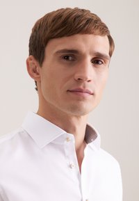 Biała koszula o teksturowanej powierzchni z usztywnionym kołnierzykiem, z kontrastującymi, wzorzystymi detalami wewnątrz kołnierzyka oraz dwoma widocznymi guzikami.