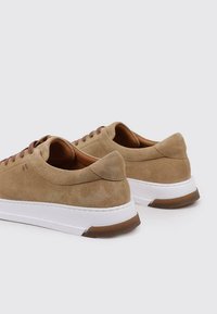 Zapatillas de ante beige con un diseño de perfil bajo. Presentan cordones marrones y una suela de goma blanca con un sutil acento marrón claro en el talón.
