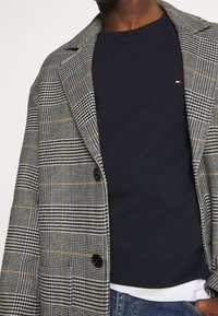 Gepunkteter Blazer in Schwarz, Weiß und Gelb mit großen Revers und zwei Knöpfen. Waffelstrukturiertes dunkelblaues Oberteil darunter. Jeans ist am unteren Rand sichtbar.