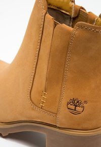 Vaaleanruskea mokkasiinibootsi, jossa on joustavat sivupaneelit, näkyvä ompelu ja puumerkki kohokuvioituna ulkopohjan kantapäässä. Sujuva pinta ja pyöristetty kärki.