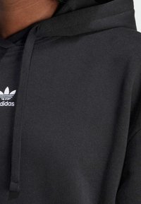 Svart hoodie i texturerad fabric med en framficka i kangaroo-stil, snörning i huvan och en vit adidas trefoil-logga på bröstet.