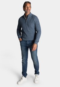 Gebreid blauw sweater met een halve ritssluiting, gecombineerd met donkerblauwe spijkerbroek en witte sneakers. Kenmerkt een gestructureerde stof.