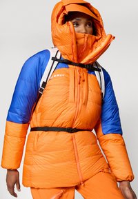 Jachetă pufosă de culoare portocalie strălucitoare cu mâneci negre, model cu carouri, guler înalt, curea ajustabilă neagră și două fermoare frontale.