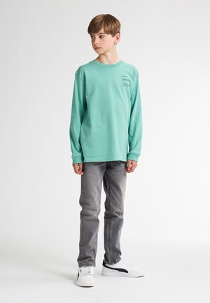 Long-sleeve mint green t-shirt with a small graphic on the chest, paired with gray jeans and white sneakers with black accents.