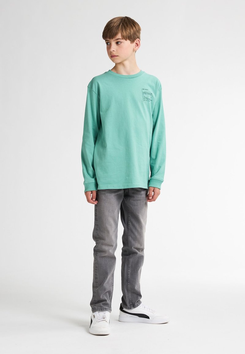 Langärmliges mintgrünes T-Shirt mit einem kleinen Grafikdesign auf der Brust, kombiniert mit grauen Jeans und weißen Sneakers mit schwarzen Akzenten.