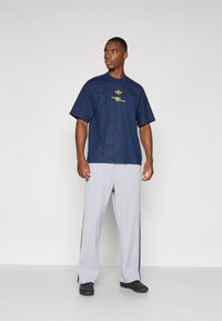 Camiseta de manga corta azul marino con un patrón texturizado y logo amarillo, combinada con pantalones gris claro con franjas laterales y zapatos negros.