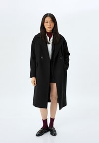 Abrigo negro con un diseño de doble botonadura, amplios solapas, bolsillos laterales y un ajuste holgado, superpuesto sobre una camisa blanca y pantalones cortos negros.