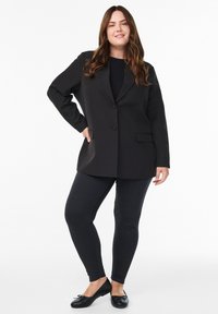 Blazer negro con un solo botón, dos bolsillos frontales y un diseño estructurado, combinado con leggings oscuros y zapatos planos negros.