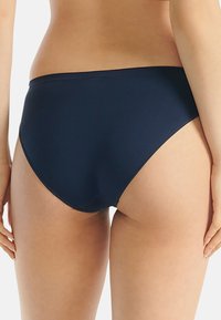 Vrouw in een eenvoudig navyblauw bikinibroekje, van achteren gezien, staand tegen een witte achtergrond.