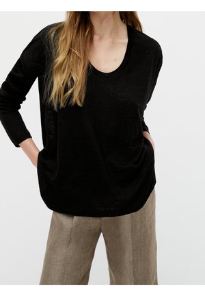 Woman wearing a loose black long-sleeve top and beige wide-leg pants with hands in pockets against a plain background.