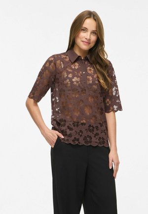 Femme aux longs cheveux bruns portant un chemisier en dentelle florale marron à manches courtes et un pantalon noir, debout avec une main dans la poche.