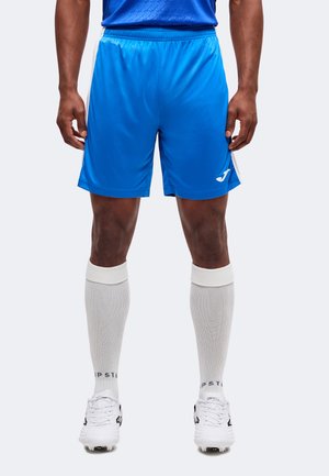 Mann trägt blaue Sportshorts, weiße knielange Socken und weiße Fußballschuhe und steht vor einem einfarbigen Hintergrund.
