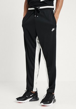 Mężczyzna w czarnych spodniach Nike z białymi paskami po bokach i czarnych butach Nike z białymi podeszwami, stoi z rękami w kieszeniach.