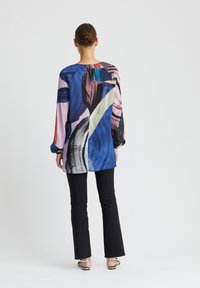 Blouse fluide avec un motif abstrait coloré en bleu, rose et vert. Manches longues et coupe ample. Associée à un pantalon noir ajusté.